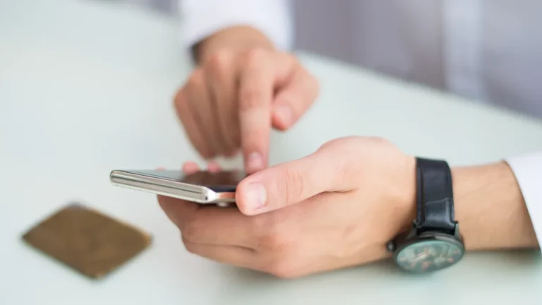Mężczyzna z zegarkiem na ręku zleca przelew w aplikacji banku na smartfonie przy biurku